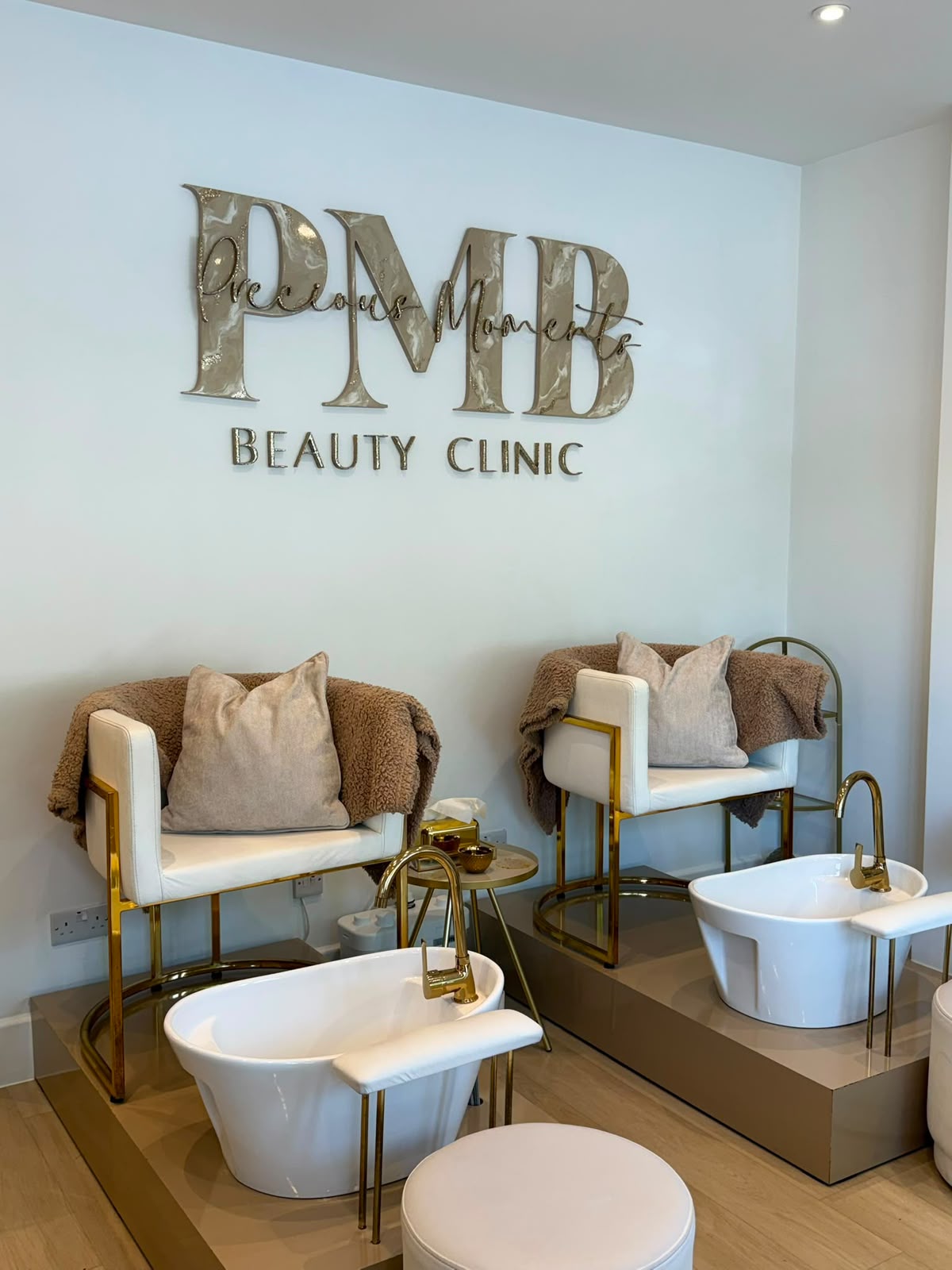 Pedicure lounge with two white and gold basin chairs under the PMB logo at Precious Moments Beauty Clinic, Banstead